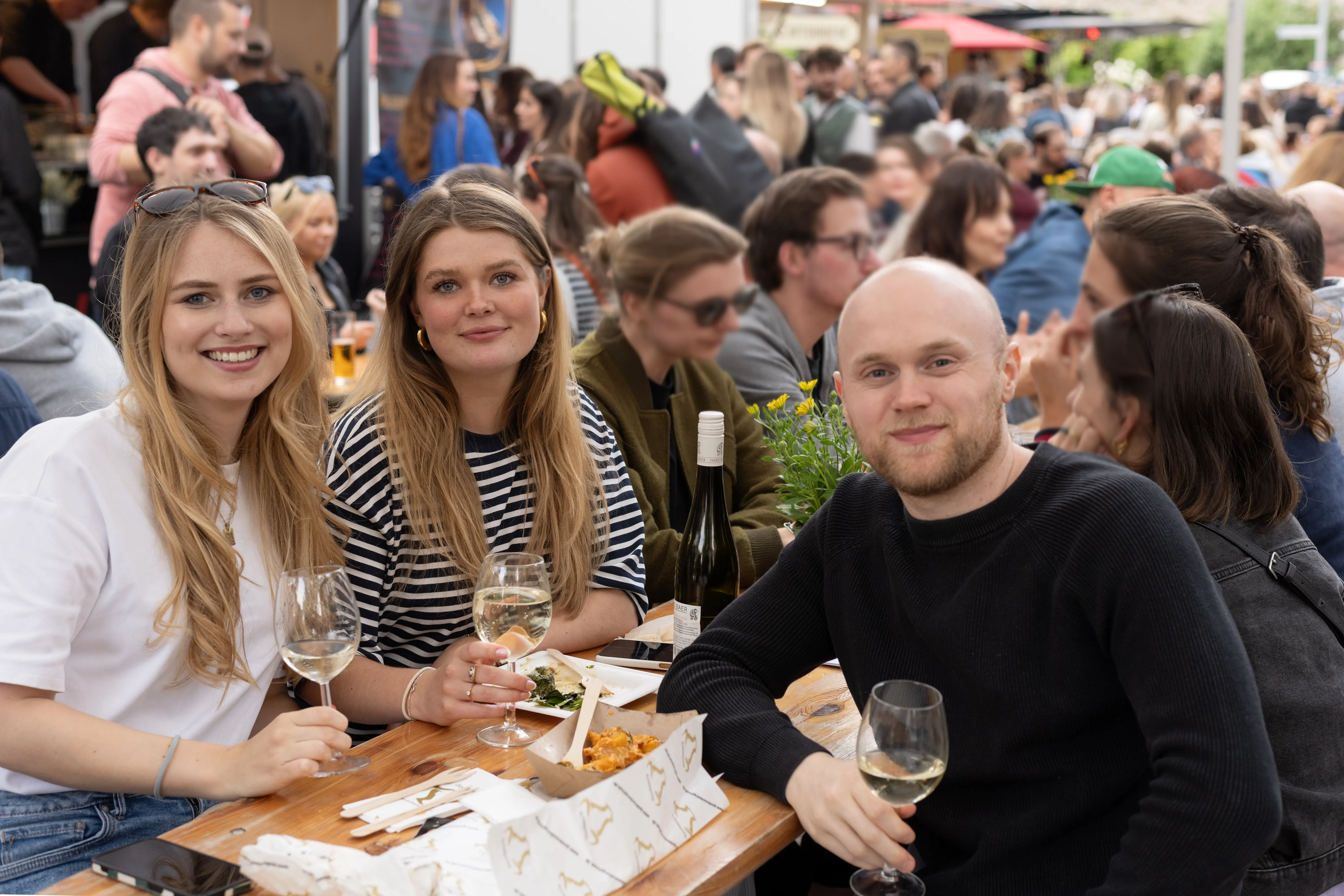 Drei Freunde genießen Wein und Streetfood auf dem Feierabendmarkt am Chlodwigplatz in Köln.