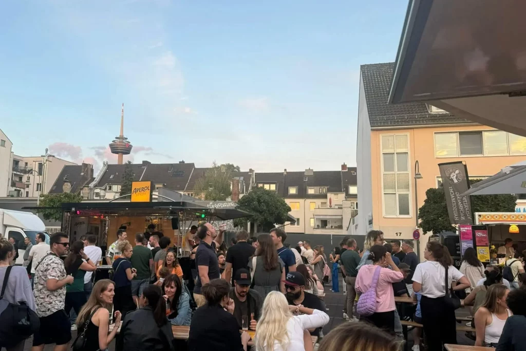 Menschen sitzen in geselliger Runde auf dem Feierabendmarkt am Neptunplatz in Köln mit Blick auf den Colonius Fernsehturm