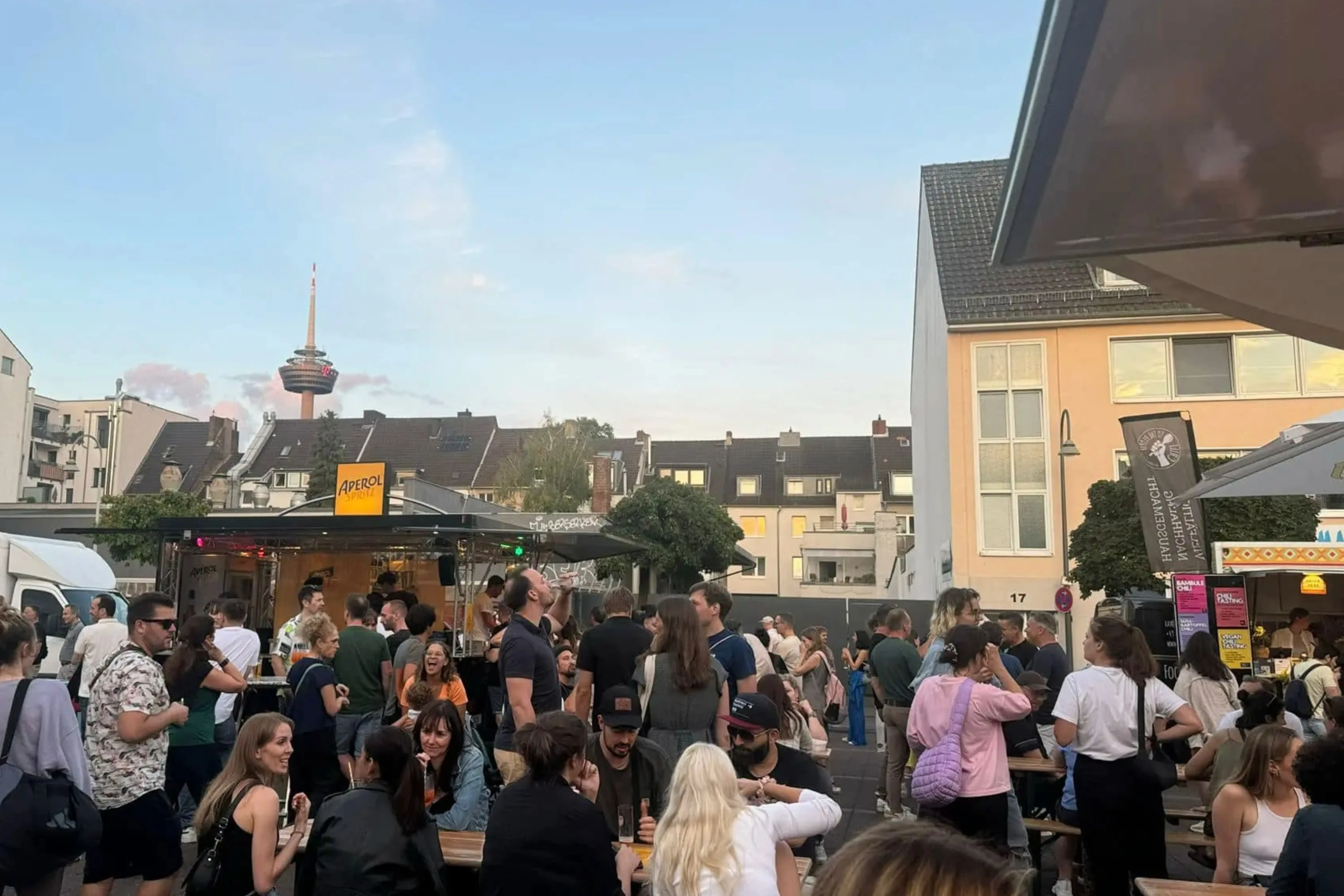 Menschen sitzen in geselliger Runde auf dem Feierabendmarkt am Neptunplatz in Köln mit Blick auf den Colonius Fernsehturm