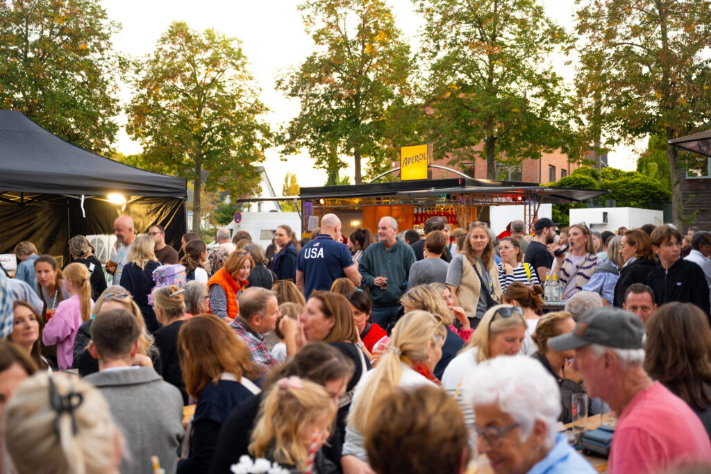 Menschenmenge in der Abenddämmerung vor dem Aperol-Stand auf dem Feierabendmarkt am Marktplatz Sürth